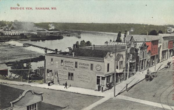 Elevated view of a row of businesses along the Fox River near the dam. There are horse-drawn vehicles in the street. Advertisements are on the side of the corner building in the foreground. Caption reads: "Bird's-Eye View, Kaukauna, Wis."