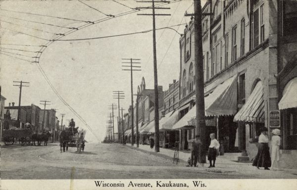 View looking up street towards the businesses along Wisconsin Avenue. There are cable car tracks running up the street, and a bank is on the corner. Pedestrians are on the sidewalk on the corner on the right, and a horse-drawn wagon is coming down the street. Caption reads: "Wisconsin Avenue, Kaukauna, Wis."