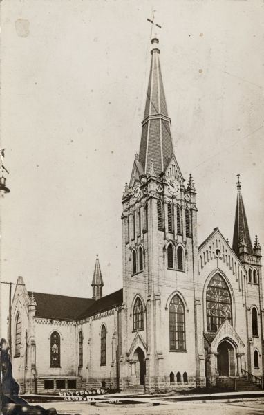 Exterior view of a gothic-style church with two steeples, and a clock tower in the taller steeple. A rose window is above the entrance. Caption reads: "Holy Cross Ch, Kaukauna, Wis."