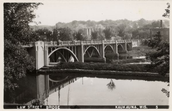 Elevated view of a bridge, with arches between the pylons, crossing the Fox River in central Kaukauna. Caption reads: "Law Street Bridge, Kaukauna, Wis."