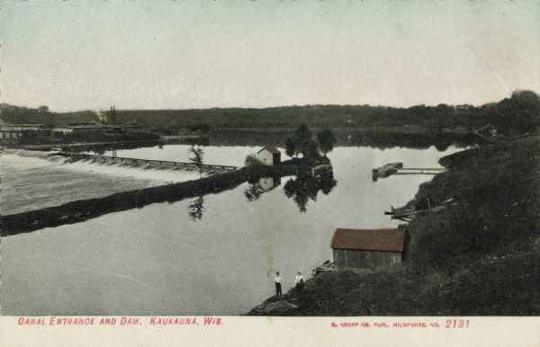 Elevated view of a canal, reservoir and dam on the Fox River. Two men are standing near a boathouse. Caption reads: "Canal Entrance and Dam, Kaukauna, Wis."