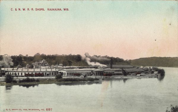 Elevated view across the Fox River towards a group of buildings next to the railroad tracks along the far shoreline. Caption reads: "C. & N. W. R. R. Shops, Kaukauna, Wis."