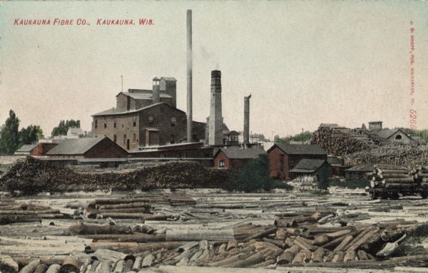 View of a fiber mill by the riverside, with a log jam in the river in the foreground. Caption reads: "Kaukauna Fibre Co., Kaukauna, Wis."
