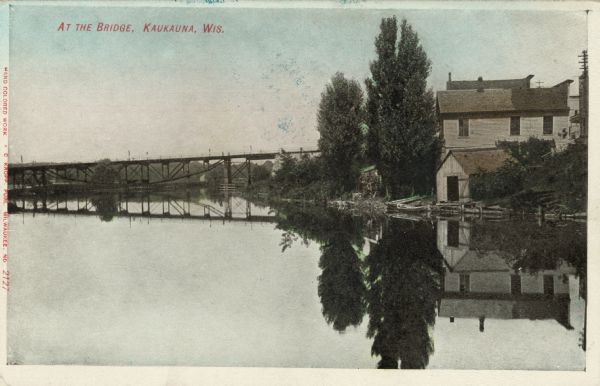 View across water towards a bridge over the Fox River. A dwelling with a boathouse is on the river bank on the right. Caption reads: "At the Bridge, Kaukauna, Wis."