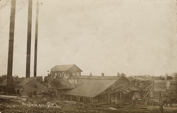 View of a sawmill. A man is standing near a horse on the right. The building in the back has many broken windows. Caption reads: "Mill Scene, Kelly, Wisconsin."