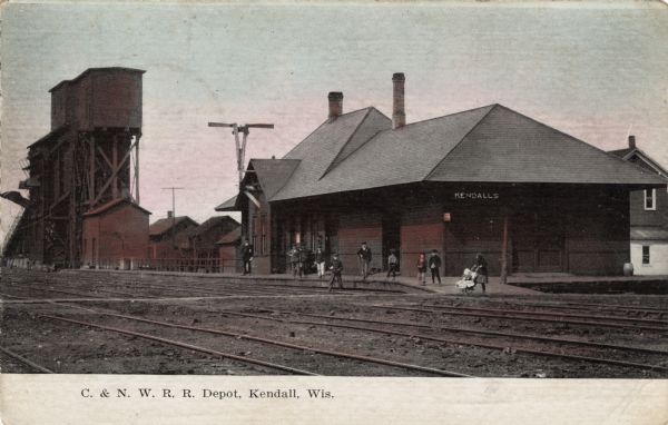 View across railroad tracks towards the depot at Kendall. Several boys are standing on the platform. Caption reads: "C. & N. W. R. R. Depot, Kendall, Wis."