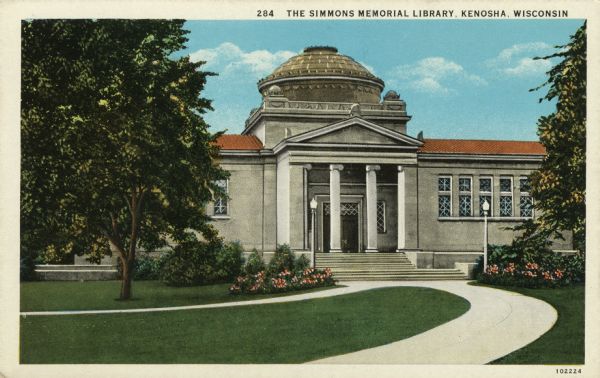 View along sidewalk toward the library with columns at the entrance and lampposts flanking the entrance. A dome is above the roof. Caption reads: "The Simmons Memorial Library, Kenosha, Wis."