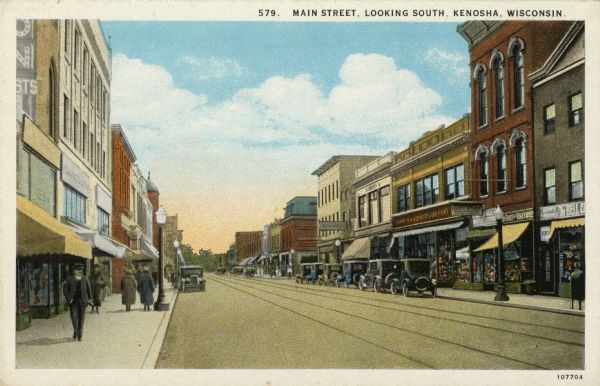 Color illustrated postcard of a street lined with shops and businesses in central Kenosha. Streetcar tracks are running down the middle of the street. Automobiles are parked along the curbs. Caption reads: "Main Street, Looking South, Kenosha, Wis."