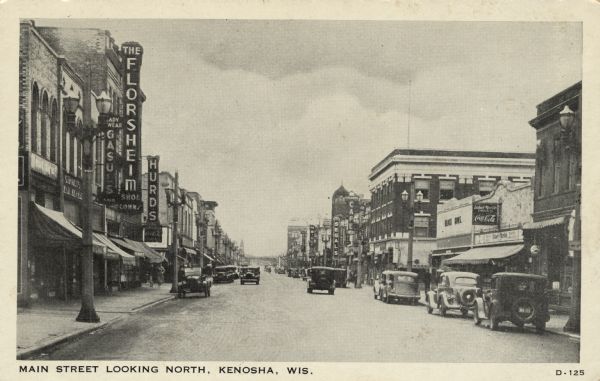 View of a central business district street lined with lampposts and electric signs. Automobiles are parked at the curbs. Caption reads: "Main Street Looking North, Kenosha, Wis."