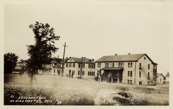 Photographic postcard view across field towards a row of apartment buildings. Caption reads: "At Keshena Falls on Highway #47, Wis."