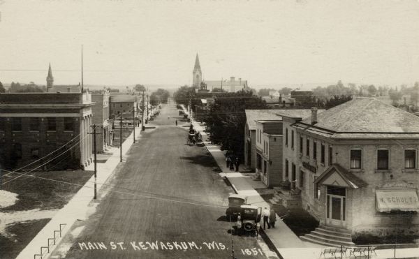 Elevated view of a central street lined with businesses. Automobiles are parked at the curbs. There is a large building with columns on the left near an open lot. Church buildings are in the distance. Caption reads: "Main St. Kewaskum, Wis."
