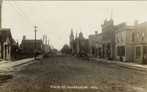 Photographic postcard view down unpaved Main Street. There is a print shop and a hotel on the left, and a dentist's office and a barber shop on the right. Horse-drawn vehicles are along the curb. Caption reads: "Main St. Kewaskum, Wis."