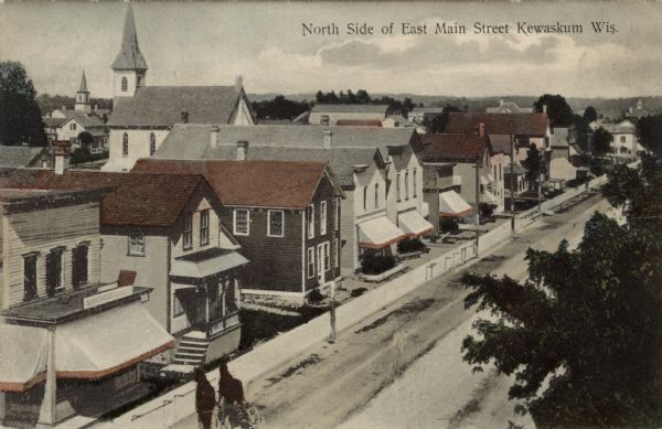 Elevated view of the central business district. A church is behind a row of businesses on the left. Hitching posts are along the sidewalk, with a horse-drawn vehicle in the left foreground. Caption reads: "North Side of East Main Street, Kewaskum, Wis."