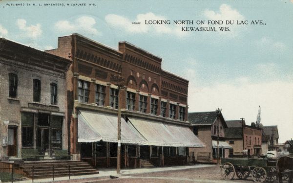 View across street towards a block lined with businesses. The Eagle Hotel on the left, and horse-drawn vehicles are on the right. Caption reads: "Looking North on Fond du Lac Ave., Kewaskum, Wis."