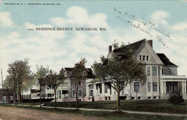 View across a tree-lined residential street, that is unpaved, towards two-story homes with porches and balconies. Caption reads: "Residence District, Kewaskum, Wis."