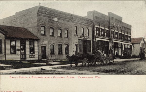 Photographic postcard view of a city block, with a store, a hotel and a few other businesses. Horses and buggies are parked at the curb. Caption reads: "Eagle Hotel and Rosenheimer's General Store, Kewaskum, Wis."