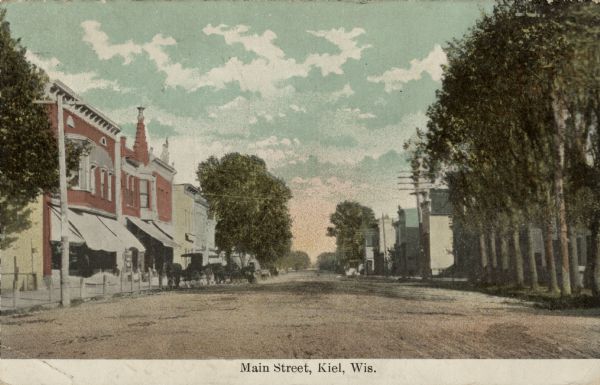 Colorized postcard view down center of unpaved Main Street. There is a block of businesses with awnings on the left. Horse-drawn vehicles are parked at hitching posts along the curb. On the right is a row of trees along the street. Caption reads: "Main Street, Kiel, Wis."
