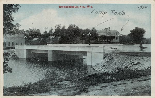 View of a bridge over the Sheboygan River. There are lampposts on each corner of the bridge, indicated by arrows. Caption reads: "Bridge Scene, Kiel, Wis."