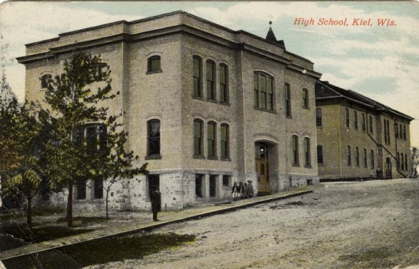 Colorized postcard view across street towards the brick high school on the left. A group of students are on the sidewalk outside of the entrance, and a man is standing on the sidewalk near the corner of the building. Caption reads: "High School, Kiel, Wis."