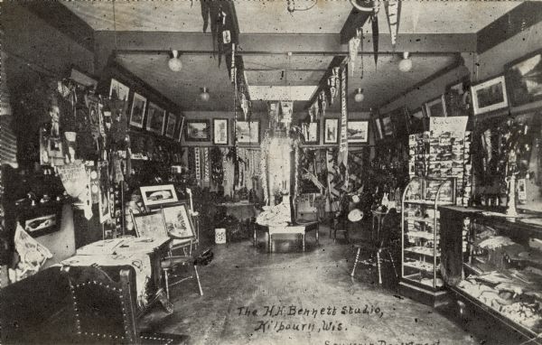 Interior view of the H.H. Bennett Studio — Souvenir Department. Framed photographs are hung along the walls, and banners are hanging from the rafters. A postcard carousel is near the glass counter on the right. Caption reads: "The H.H. Bennett Studio, Kilbourn, Wis."