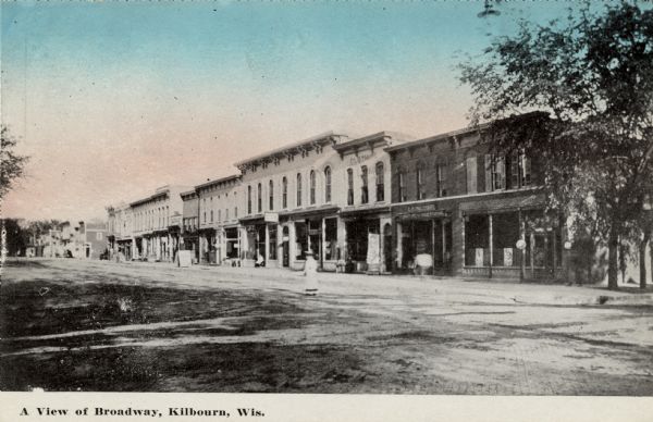 View across street towards the businesses lining the block. A woman is walking in the street.Caption reads: "A View of Broadway, Kilbourn, Wis."