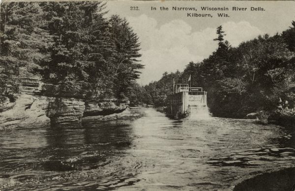 View across river towards the rear of the excursion boat "Apollo" in the narrows of the dells. Rock formations are on the left. Caption reads: "In the Narrows, Wisconsin River Dells, Kilbourn, Wis."
