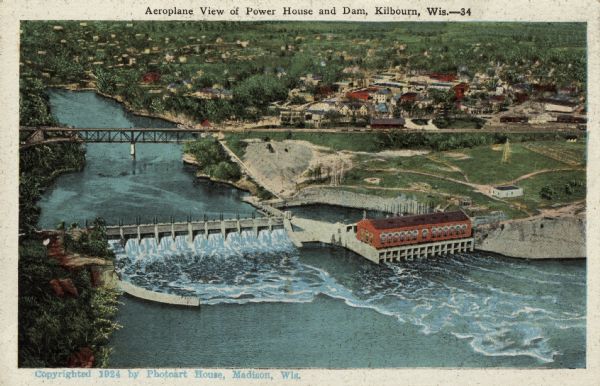 Illustration based on an aerial photograph of a powerhouse and dam on the Wisconsin River. A railroad bridge is upriver. The city of Kilbourn (now Wisconsin Dells) is just beyond the railroad tracks. Caption reads: "Aeroplane View of Power House and Dam, Kilbourn, Wis."