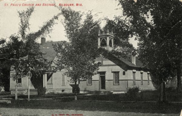 View across street towards a small wooden church with a bell tower, and neighboring rectory on the left. Caption reads: "St. Paul's Church and Rectory, Kilbourn, Wis."