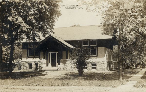 Sepia-toned photographic postcard of a one-story library with stone foundation and a large gable over the entrance. Caption reads: "Public Library, Kilbourn, Wis."