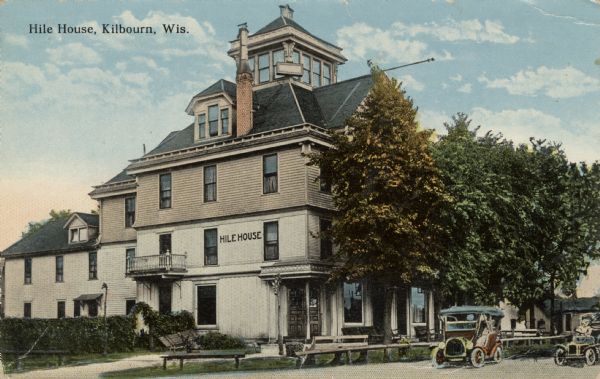 Hand-colored postcard of the Hile House hotel. Two automobiles are parked on the street on the right. Caption reads: "Hile House, Kilbourn, Wis."