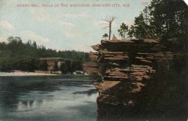 Illustration of a bluff at the Dells, with a woman is sitting on the top near a tree. There are bluffs on the other side of the river in the background.Caption reads: "Hawk's Bill, Dells of the Wisconsin, Kilbourn City, Wis."
