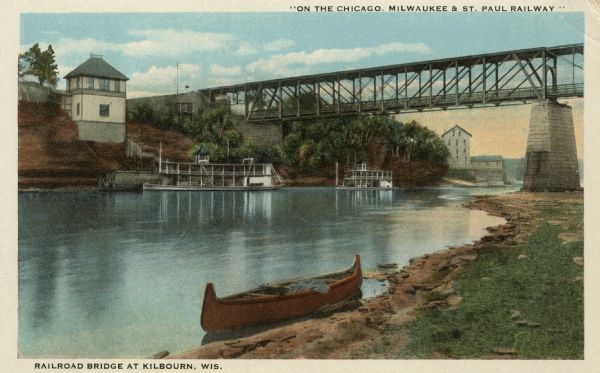 Colorized postcard view of the railroad bridge over the Wisconsin River at Kilbourn. A canoe is along the shoreline in the foreground, and excursion boats are along the far shoreline. Caption at bottom reads: "Railroad Bridge at Kilbourn, Wis."<p>Kilbourn City was founded in 1857 by Byron Kilbourn, the name was changed in 1931 to Wisconsin Dells.</p>