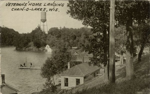 Sepia-toned photographic postcard view from hill looking down towards men standing on the pier at the Veterans' Home. A water tower is across the water. Caption reads: "Veterans' Home Landing, Chain-O-Lakes, Wis."