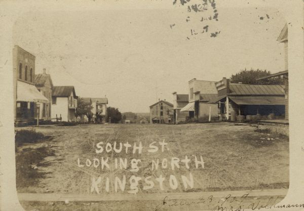 View down an unpaved street lined with businesses. Caption reads: "South St. Looking North, Kingston."