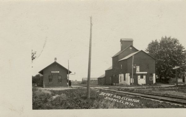 View towards a railroad crossing, with the depot on the left side of the tracks, and a grain elevator on the right. A sign along the road reads: "Look Out For The Cars." Caption reads: "Depot and Elevator, Knowles, Wis."