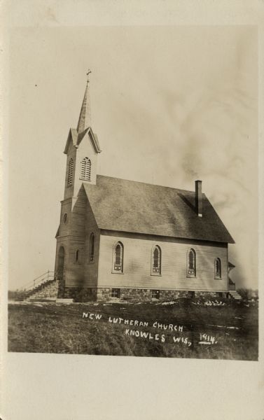 View towards a wooden church with a stone foundation, steeple and Gothic arched stained glass windows. The stone foundation extends to support the steep front steps to the arched entrance, and a small porch at the back of the church has steps to the side yard on the right, which has piles of dirt and a wheelbarrow. Caption reads: "New Lutheran Church, Knowles, Wis."