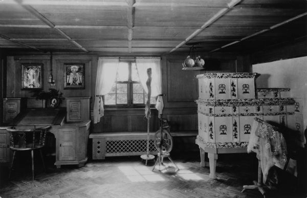Photographic postcard view of the Waelderhaus work room. There is a desk on the left, with two framed prints above it. There is an Austrian Kachelofen (tile stove) on the right. A spinning wheel and work bench are underneath the window. Caption on back reads: "Werkzimmer (workroom) in Waelderhaus, the Girl Scout House of Kohler, Wisconsin, U.S.A. Note Austrian Kachelofen (tile stove)."