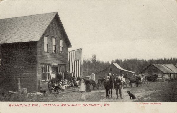 Several people are gathered on the front stoop of a building, probably a general store. Horses and wagons are in the road. Log buildings are in the background.