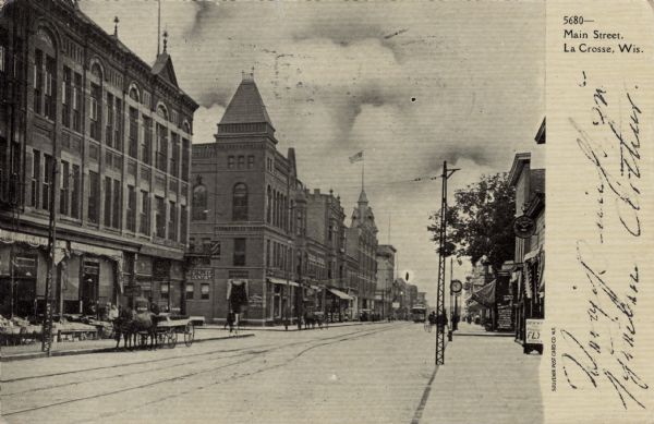 View down sidewalk towards the businesses along Main Street. A streetcar is coming down tracks running down the center of the street, and there are horse-drawn vehicles. Caption reads: "Main Street, La Crosse, Wis."