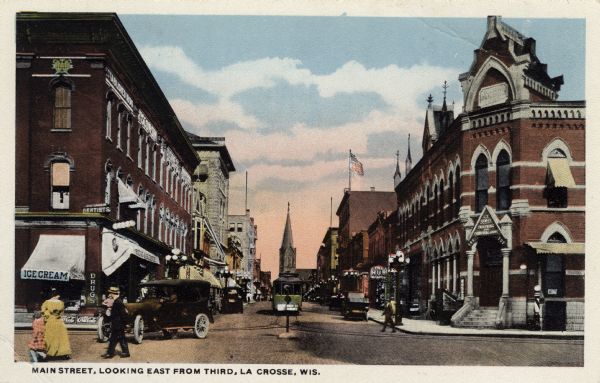 View of a central business district. A streetcar and automobiles are in the street. Street lamps are along the curbs, and pedestrians are walking on the street and sidewalks. Caption reads: "Main Street, looking east from Third, La Crosse, Wis."