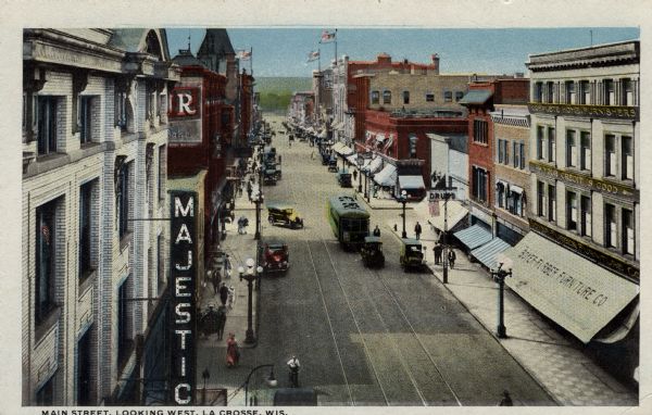 Elevated view of a central business district. The Majestic theater is on the left, and a furniture store is on the right. A street car, pedestrians and automobiles are in the street. Caption reads: "Main Street, Looking West, La Crosse, Wis."