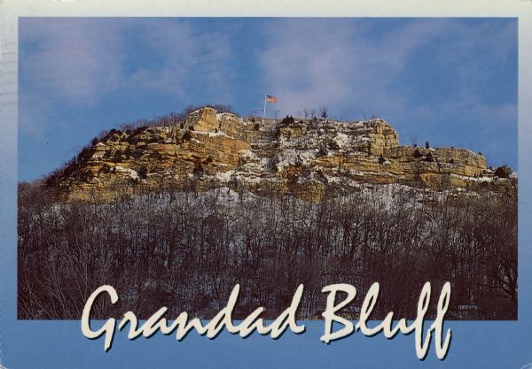 View of Grandad Bluff with snow on the ground. There is an American Flag flying from a flagpole on the top of the bluff. Text on back of postcard reads: "Feel winter's cold northwest winds from the observation point on top of Grandad Bluff. On a clear day you can see La Crosse, the Mississippi River, the Minnesota river bluffs and Iowa, downriver. Grandad Bluff, at 590 feet, is the highest point in La Crosse County."