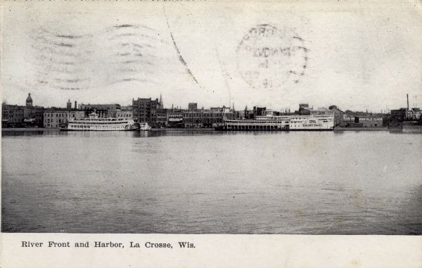 View across the Mississippi River towards the city of La Crosse. Two riverboats are at the river's edge. Caption reads: "River Front and Harbor, La Crosse, Wis."
