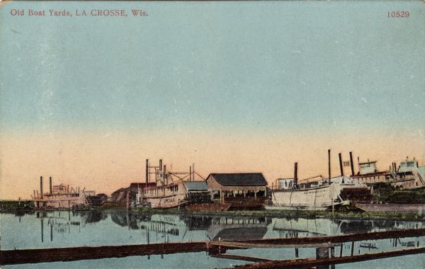 Colorized view of a boat yard on the Mississippi River. Riverboats are moored along the shoreline. Caption reads: "Old Boat Yards, La Crosse, Wis."