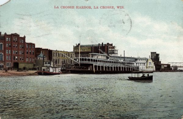 View across water towards the La Crosse Harbor. On the right are two people in a small excursion boat. Along the shoreline is the paddle steamer "St. Paul," and a small tugboat is on the left. Caption reads: "La Crosse Harbor, La Crosse, Wis."