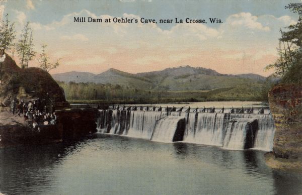 Elevated view upriver toward a dam in a river, with hills in the background. A group of people are on the elevated bank on the left. Caption reads: "Mill Dam at Oehler's Cave near La Crosse, Wis."