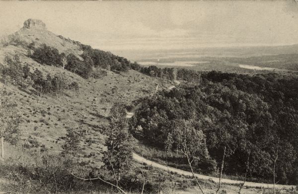 View towards a hill, on the far left in the distance, with a geologic formation at the top. A road is winding through the hillside. The Mississippi River is in the distance.