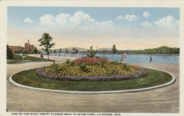 Hand-colored view of a circular flower bed next to the river, with an interstate bridge in the distance. Caption reads: "One of the Many Pretty Flower Beds in Levee Park, La Crosse, Wis."