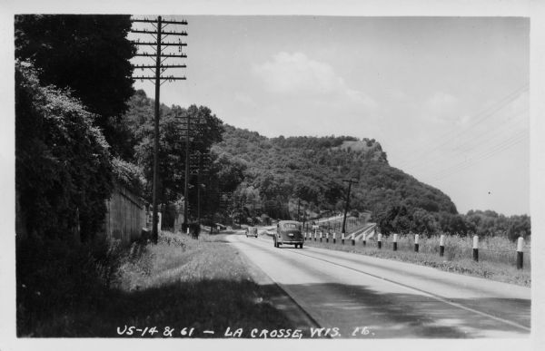 Photographic postcard view of a two-lane Interstate highway along railroad tracks. Two automobiles are moving down the road in the right lane, heading towards a bluff. Caption reads: "U.S. 14 & 61 — La Crosse, Wis."