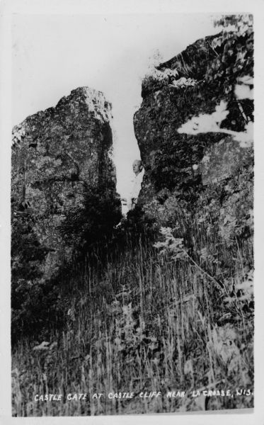 Photographic postcard view of a rock formation on a hillside, with a man standing in between looking towards the left. Caption reads: "Castle Gate at Castle Cliff Near La Crosse, Wis."